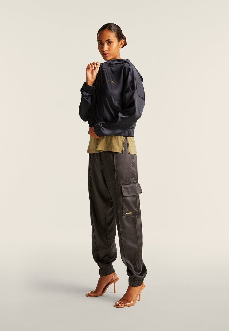 Chaqueta con capucha azul marino y puños elásticos, camisa verde oliva de capas y pantalones cargo negros de satén con bolsillos, combinados con tacones marrones de tiras.