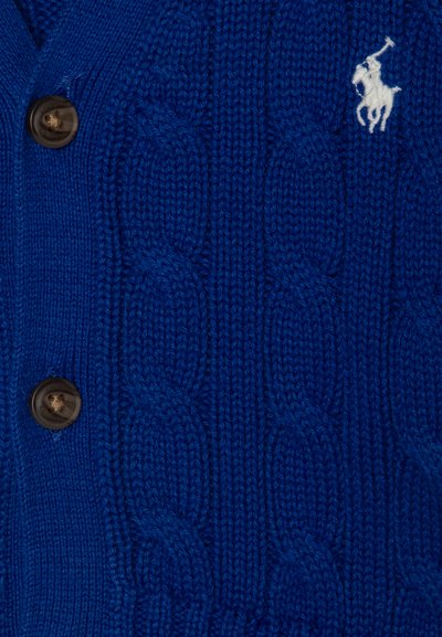 Blau gestrickte Wolljacke mit Kabelstrickmuster, vier braunen Knöpfen und einem kleinen weißen Logo eines Pferdes mit einem Reiter auf der Brust.