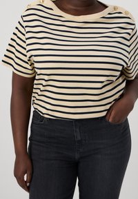 Cream and navy striped t-shirt with short sleeves, round neckline, and two decorative buttons on the shoulder. Paired with dark jeans.