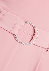 Tissu rose à surface texturée, avec une large ceinture maintenue par une boucle circulaire en argent. Détails de plis doux près de la boucle.