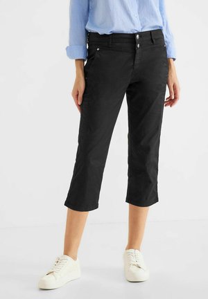 Femme portant un pantalon court noir à double fermeture boutonnée, une chemise bleu clair et des baskets blanches, debout devant un fond blanc uni.