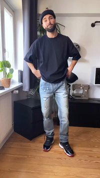 Homem a usar um boina preta, t-shirt preta oversized, jeans claros rasgados e ténis pretos, de pé numa sala iluminada com uma máquina de costura e plantas.
