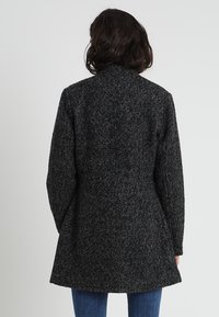 Kvinna med axellångt mörkt hår bär en lång, mörkgrå texturerad kappa och blå jeans, vänd bort från kameran mot en enfärgad bakgrund.