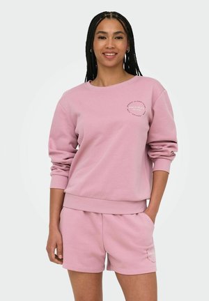 Ung kvinde med flettet hår bærer matchende lyse lilla sweatshirt og shorts, smilende med den ene hånd i lommen på en ensfarvet baggrund.