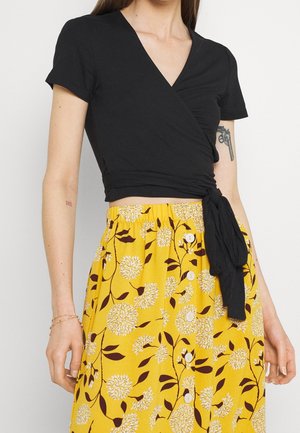 Schwarzes Wickeloberteil mit kurzen Ärmeln und gerafften Details, kombiniert mit einem gelben Rock mit Blumenmuster und Knöpfen vorne.