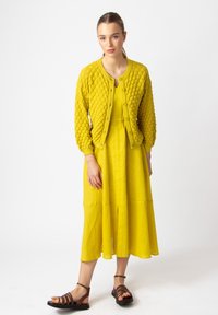 Kollane tekstureeritud kardigan ümmarguse kaelusega, kombineeritud sobiva kollase kleidiga, millel on põlvekohale ulatuv seelik. Pruunid rihmadega sandaalid.