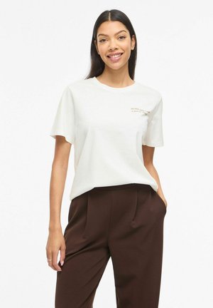 Femme souriante aux longs cheveux foncés porte un t-shirt blanc à manches courtes avec un petit texte et un pantalon marron taille haute, main dans la poche.