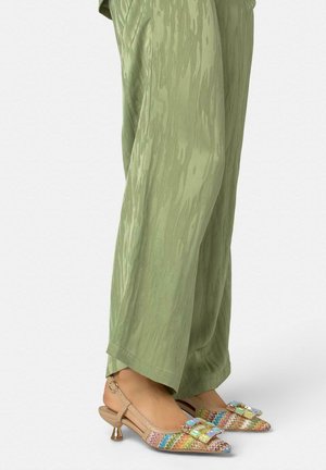 Pies con zapatos slingback tejidos multicolores de tacón bajo y grandes hebillas decorativas, combinados con pantalones sueltos de textura ligera en color verde claro.