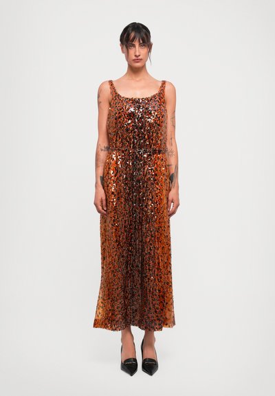 Femme portant une robe midi sans manches à imprimé léopard orange et noir ornée de sequins, avec des escarpins noirs à bout pointu, se tenant contre un fond uni.