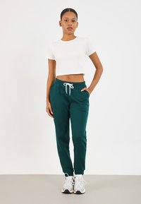 Tyrkysové joggers nohavice s bielou nastaviteľnou šnúrkou, so bočnými vreckami. V kombinácii s priliehavým bielym cropped tričkom a sivými teniskami.