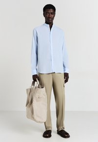 Camicia azzurra chiara a maniche lunghe con colletto a bard, pantaloni beige e una borsa a tote in tela beige con tasche laterali, su uno sfondo semplice.