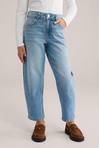 Isik, kes kannab hele siniseid teksapükse väike hõbedaste neetidega ja pruune seemisnahast loafereid, käsi taskus ja valget pika varrukaga toppi.