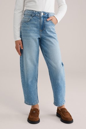 Osoba ubrana w jasnoniebieskie dżinsy z małymi srebrnymi ćwiekami i brązowe zamszowe mokasyny, ręka w kieszeni, biała bluzka z długim rękawem.