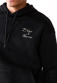 Sudadera negra con bolsillo frontal, con bordado en blanco del texto "Naturaleza en Armonía" y dos siluetas de pájaros. Textura de tela suave.