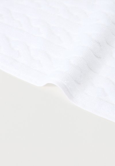 Λευκό υφασμάτινο πάπλωμα με κυματιστή υφή και καμπύλη άκρη πάνω σε απλό ανοιχτόχρωμο φόντο.