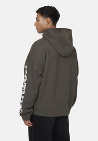 Sweat à capuche vert olive, en coton, avec une coupe décontractée et un texte graphique blanc vertical sur la manche gauche.