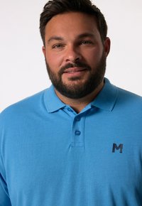 Světle modré polo tričko s límečkem, třemi knoflíky a černým logem na levém hrudi; texturovaná látka, volný střih.