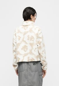 Άτομο με κοντά σκούρα μαλλιά που φοράει μπεζ και λευκό φλις μπουφάν με σχέδιο και γκρι παντελόνι εργασίας, βλέπει προς τα πίσω με φόντο ένα απλό φόντο.