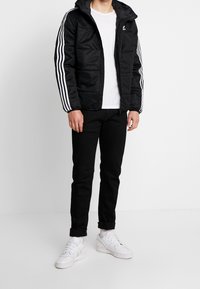 Svart vadderad jacka med huva och vit tre-randsdesign på ärmarna, tillsammans med svarta jeans och vita sneakers.