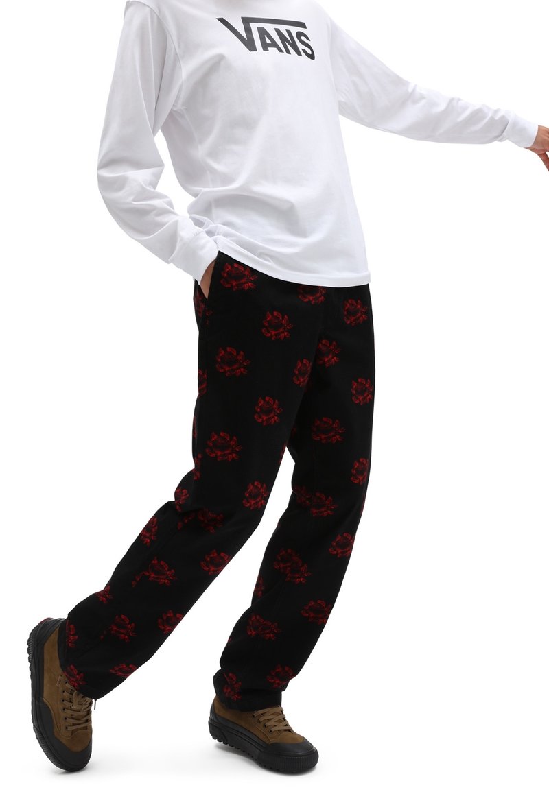 Pantalon noir avec un motif floral rouge, associé à une chemise blanche à manches longues et des baskets marron à semelles noires.
