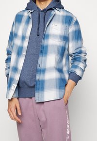 Personne portant une chemise à carreaux bleue boutonnée par-dessus un sweat à capuche bleu marine et un pantalon de survêtement rose avec un texte blanc vertical sur la jambe gauche.
