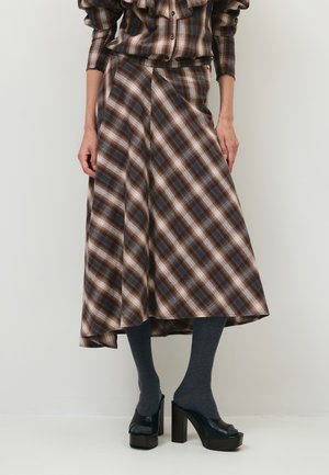 Vrouw draagt een bruine geruite midirok met bijpassende top, donkergrijze panty's en zwarte plateauhoge hakken, staande tegen een effen achtergrond.