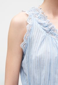 Αμάνικο ανοιχτό μπλε μπλουζάκι με λεπτές κάθετες ρίγες και δαντέλα με τρύπες γύρω από τη μασχάλη και το ντεκολτέ, φορεμένο με κολιέ με χάντρες.