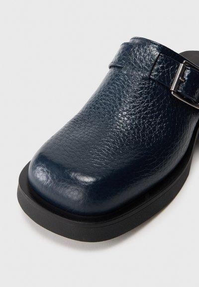 Zueco de cuero azul marino con acabado texturizado, punta redondeada, hebilla metálica ajustable y suela de goma negra.