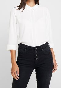 Blusa bianca con colletto, maniche 3/4 e chiusura con bottoni. Abbinata a jeans neri in denim a vita alta con cinque bottoni.