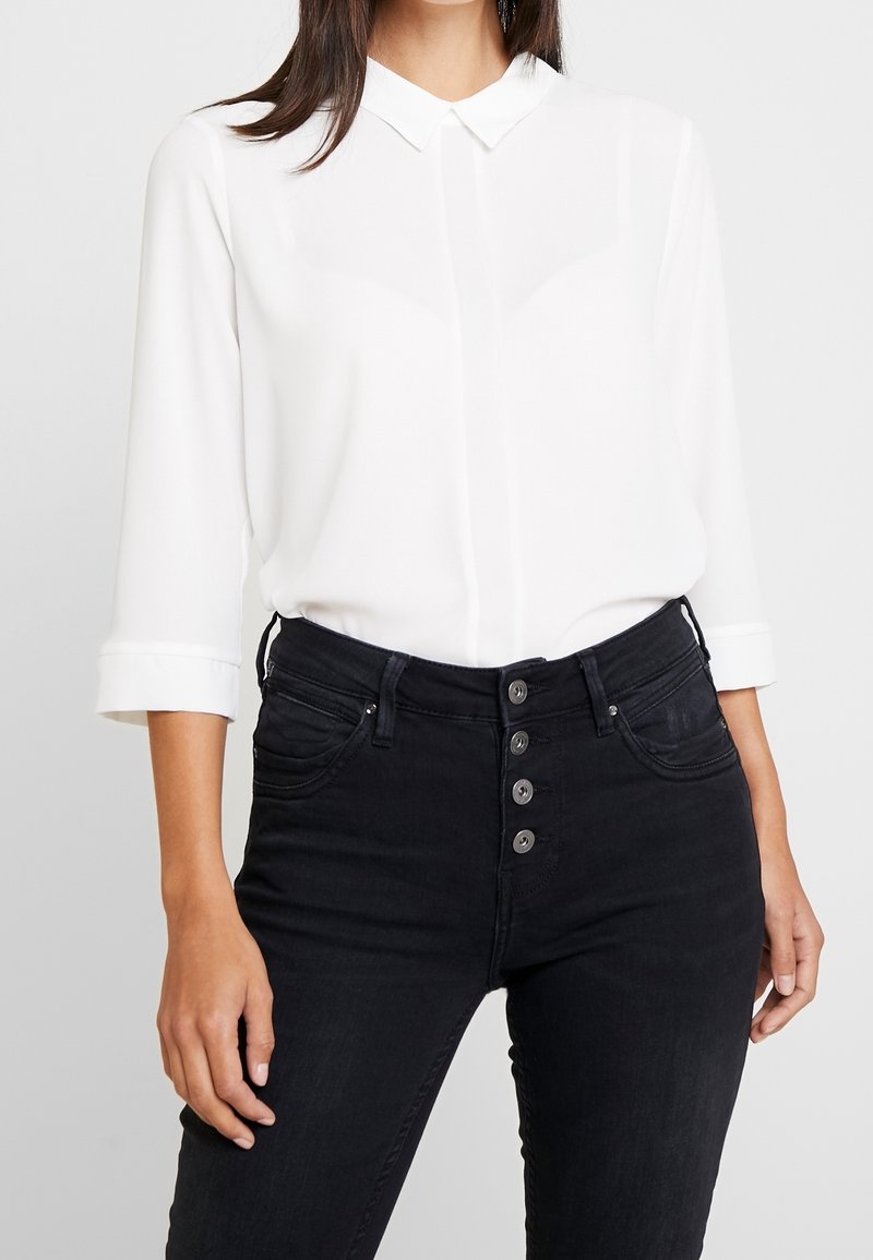 Blusa bianca con colletto, maniche 3/4 e chiusura con bottoni. Abbinata a jeans neri in denim a vita alta con cinque bottoni.