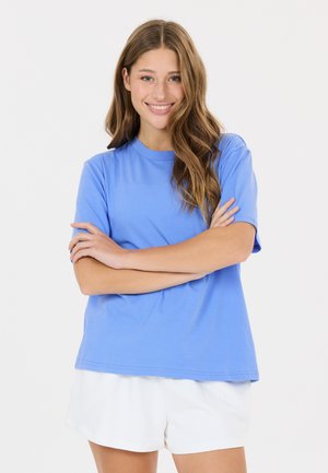 Jeune femme aux longs cheveux ondulés souriante, portant une chemise bleue à manches courtes et un short blanc, bras croisés devant un fond blanc.