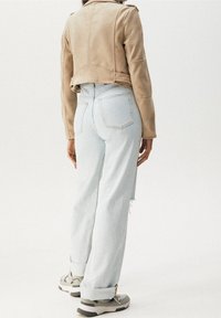 Veste courte en daim couleur fauve, jean taille haute bleu clair, et baskets grises, avec poches arrière visibles et ourlets légèrement effilochés.