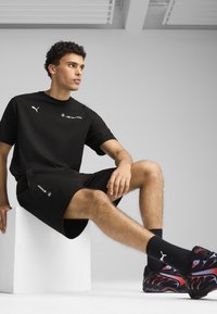 Sort Puma t-shirt og shorts med BMW Motorsport logo. Modellen har mønstrede sneakers og sorte ankelstrømper på, mens han sidder på en hvid blok.