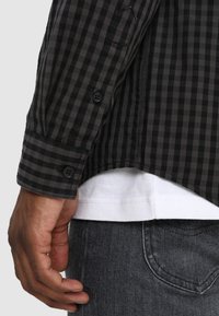 Mano maschile accanto a una camicia a maniche lunghe a scacchi neri e grigi sopra una maglietta bianca e jeans in denim grigi.
