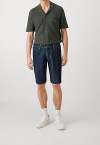 Camisa de manga corta, de color verde oscuro, con cuello y un bolsillo, combinada con pantalones cortos de mezclilla oscuros. El calzado incluye zapatillas blancas y calcetines.