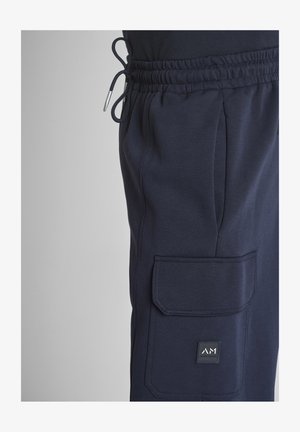 Pantalon de survêtement bleu marine avec ceinture élastique, cordon de serrage, et poche rabat ornée d'un petit patch carré avec le logo de la marque.