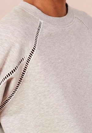 Sudadera gris claro con un patrón decorativo de punto abierto a lo largo de la costura del hombro y el brazo, usada por una persona de tono de piel medio.