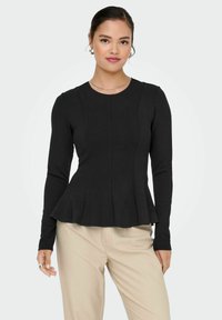 Black long-sleeve top with a fitted bodice and flared peplum hem. Round neckline, textured fabric, paired with light-colored pants.