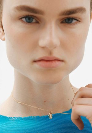 Collar de oro con un colgante de destello solar, llevado sobre un suéter azul con un escote deshilachado, que presenta una cadena suave y delicada.