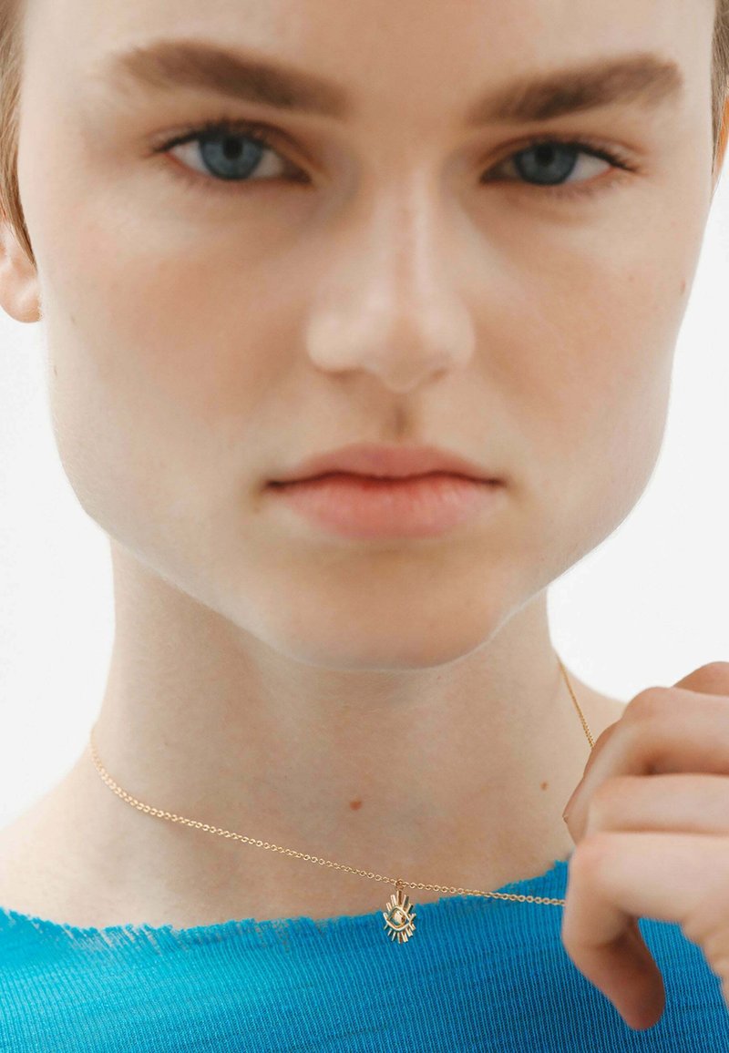 Collar de oro con un colgante de destello solar, llevado sobre un suéter azul con un escote deshilachado, que presenta una cadena suave y delicada.