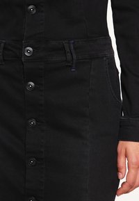 Svart denimklänning med knappstängning fram, sidofickor och justerbar midja. Har en slät textur och en figurnära silhuett.