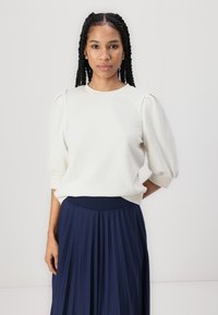 Cream-colored sweater with puffed sleeves, soft texture, and round neckline, paired with a navy pleated skirt featuring an elastic waistband.