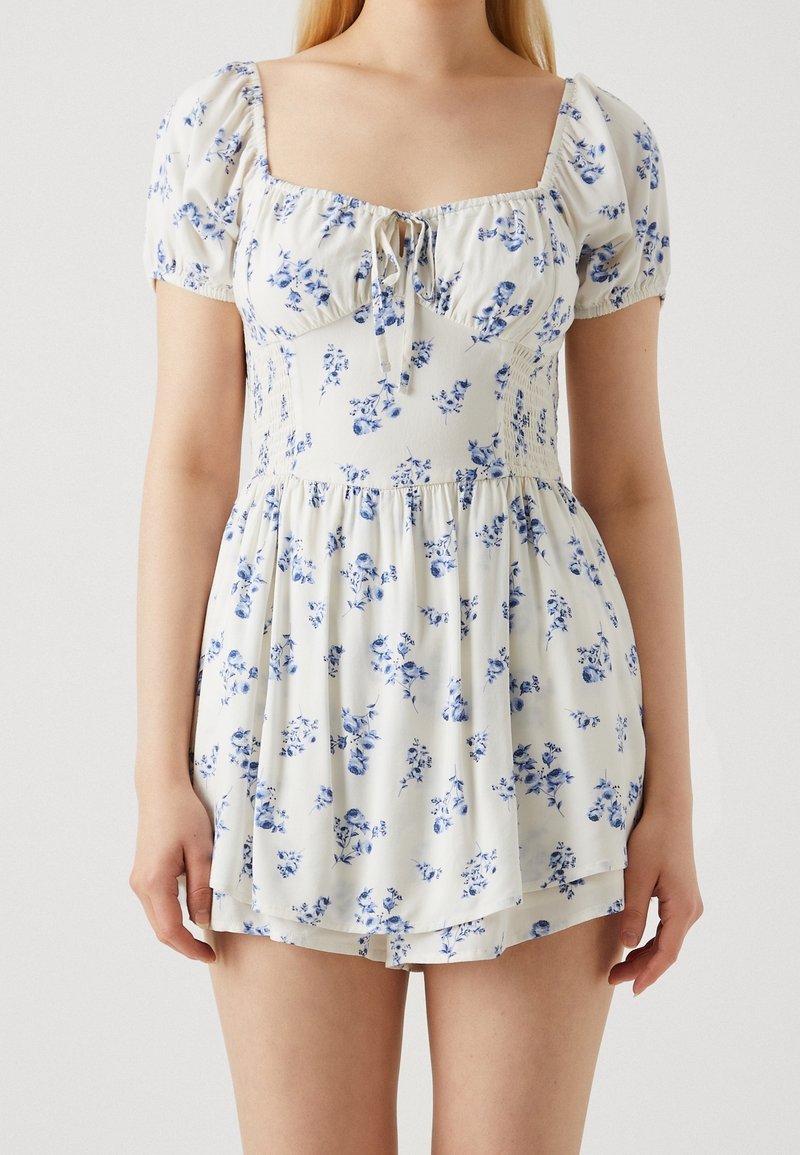 Femme portant une robe blanche à manches courtes avec un imprimé floral bleu, un décolleté carré et une jupe froncée.