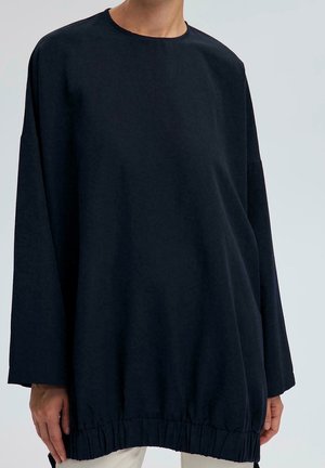 Person trägt ein locker sitzendes langärmliges schwarzes Shirt mit rundem Ausschnitt und gerafftem Saum, dargestellt vom Hals bis zur Mitte des Oberschenkels.