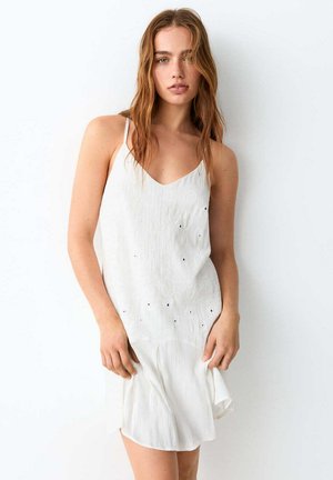 Young woman with loose wavy hair wearing a sleeveless white dress with embroidered eyelet details, standing against a plain white background.