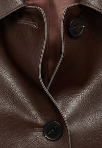 Brązowa skórzana kurtka z wyraźnym kołnierzem, dwoma czarnymi guzikami i kontrastującym beżowym przeszyciem wzdłuż krawędzi listwy guzików.