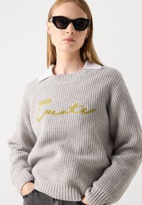 Gray knitted sweater with ribbed texture, featuring a gold embroidered "C." Collared white shirt visible underneath. Black cat-eye sunglasses.