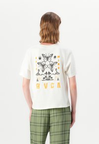 White short-sleeve t-shirt featuring a black and yellow butterfly and botanical graphic on the back, paired with green plaid pants.