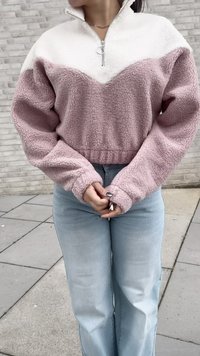 Donna che indossa un pullover in pile rosa e bianco con colletto con zip e jeans azzurri, in piedi contro uno sfondo di parete grigia piastrellata.