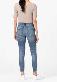 Kvinna i ljusbrun kortärmad topp, blå högmidjade skinny jeans och vita sneakers, som står vänd bort mot en vit bakgrund.